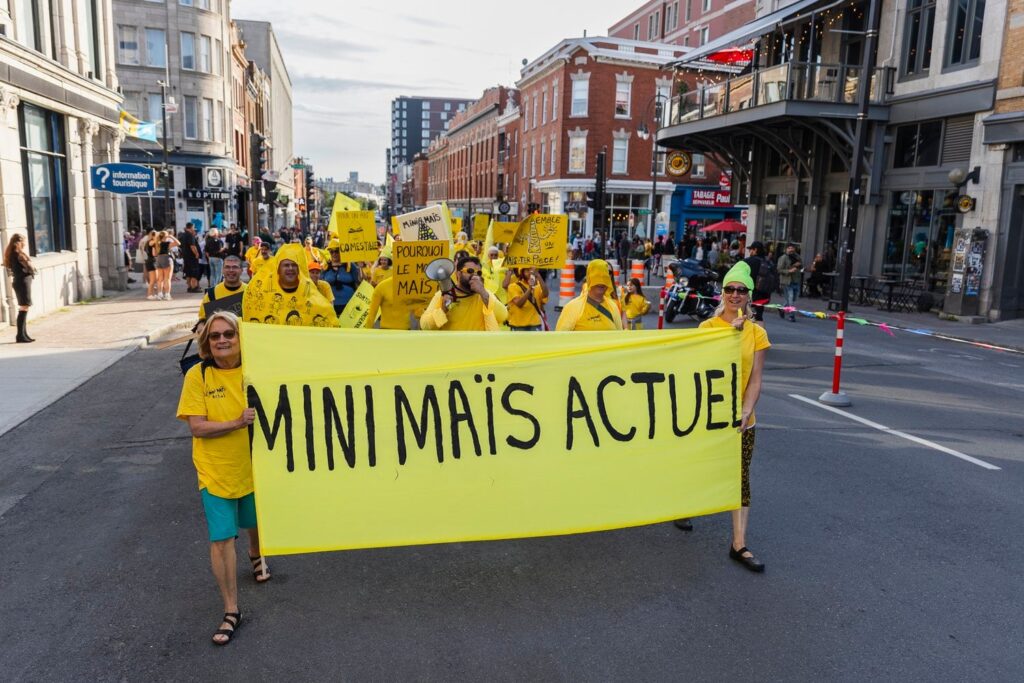 Amas de gens dans la rue habillé-es de jaune et portant un banderole écrit Mini Maïs Actuel.