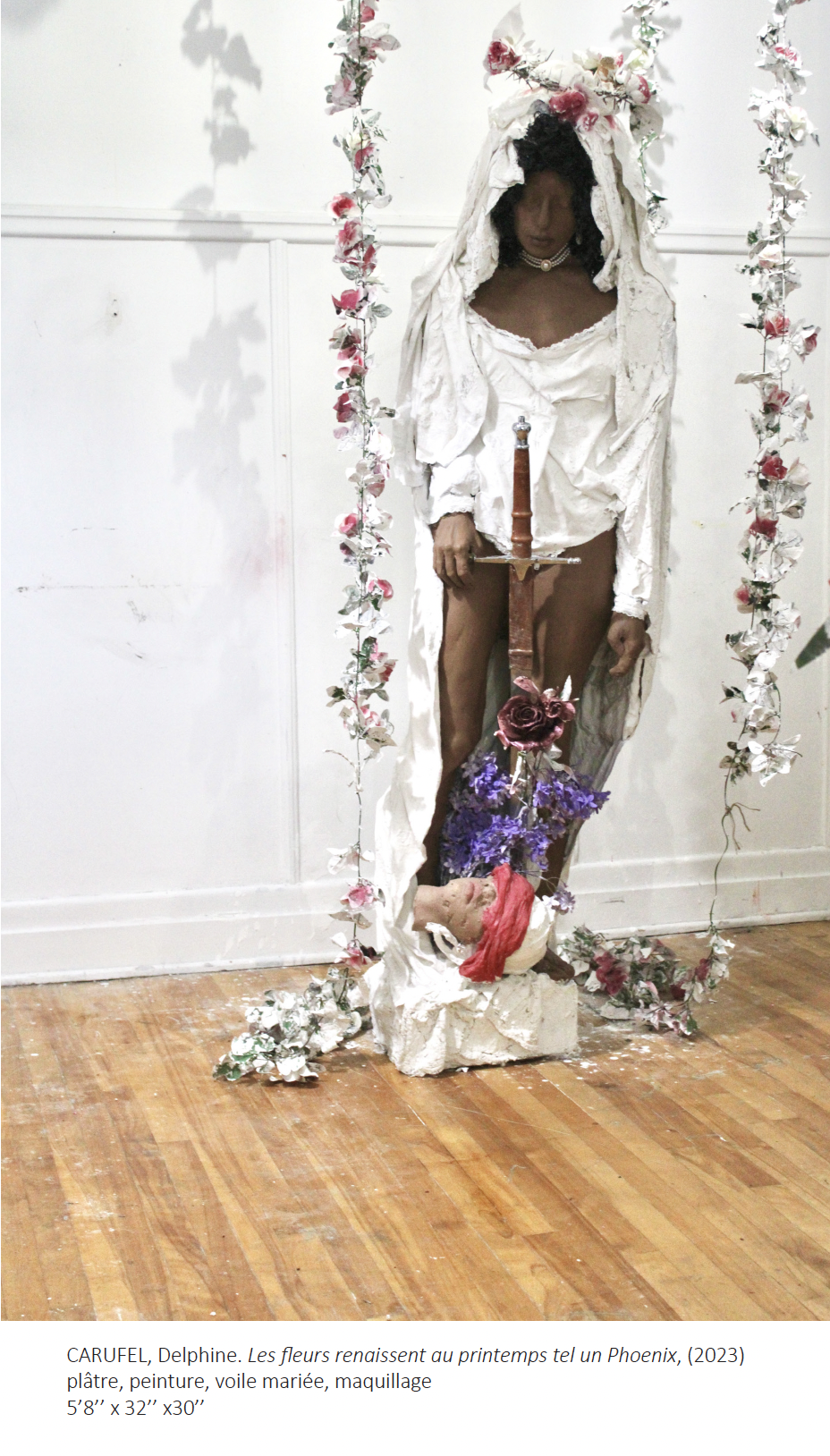 Sculpture grandeur nature d'un corps humain à la peau brune foncée, regardant au sol, portant une robe et coiffe blanche avec fleurs.