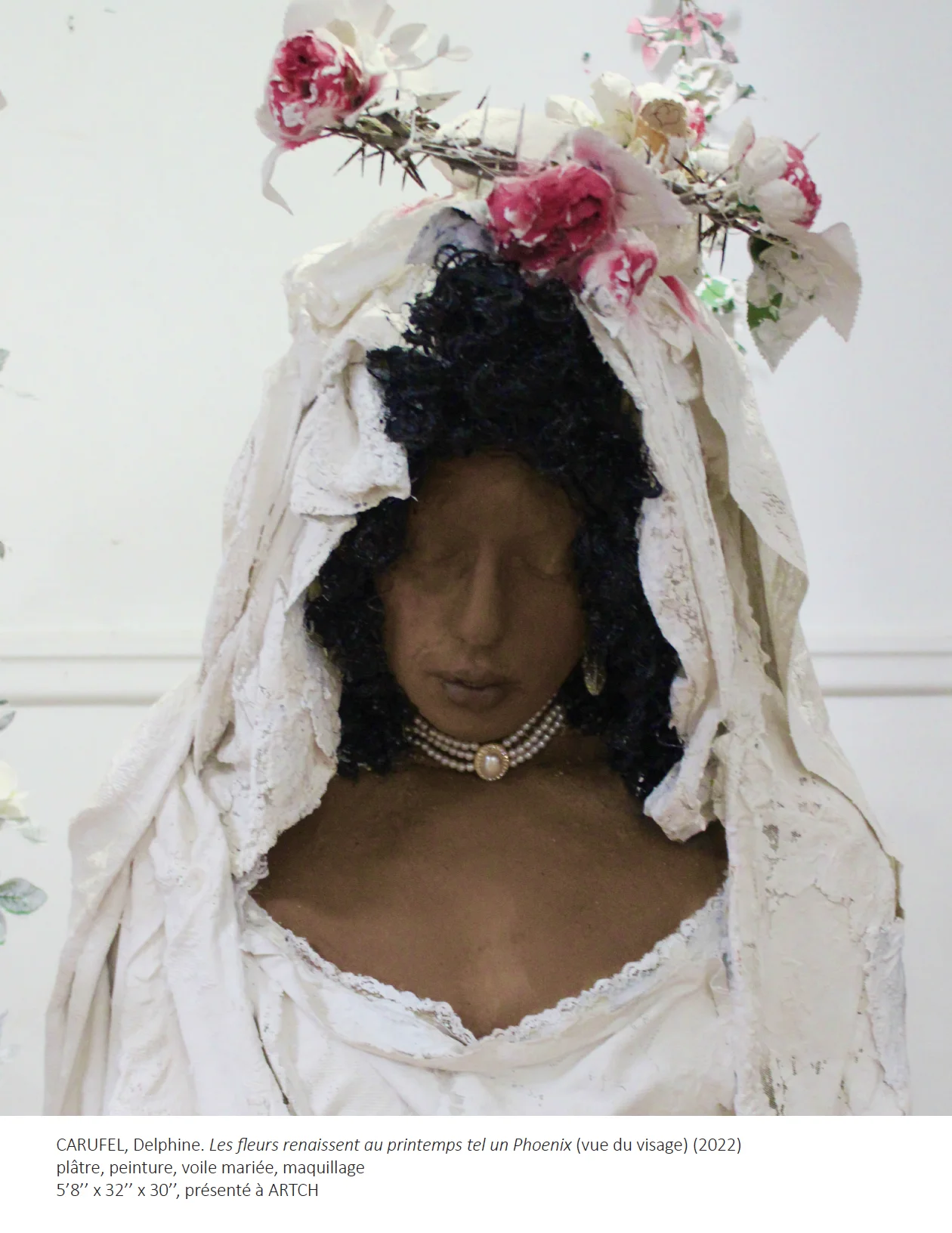 Détail du visage de la Sculpture grandeur nature d'un corps humain à la peau brune foncée, regardant au sol, portant une robe et coiffe blanche avec fleurs.