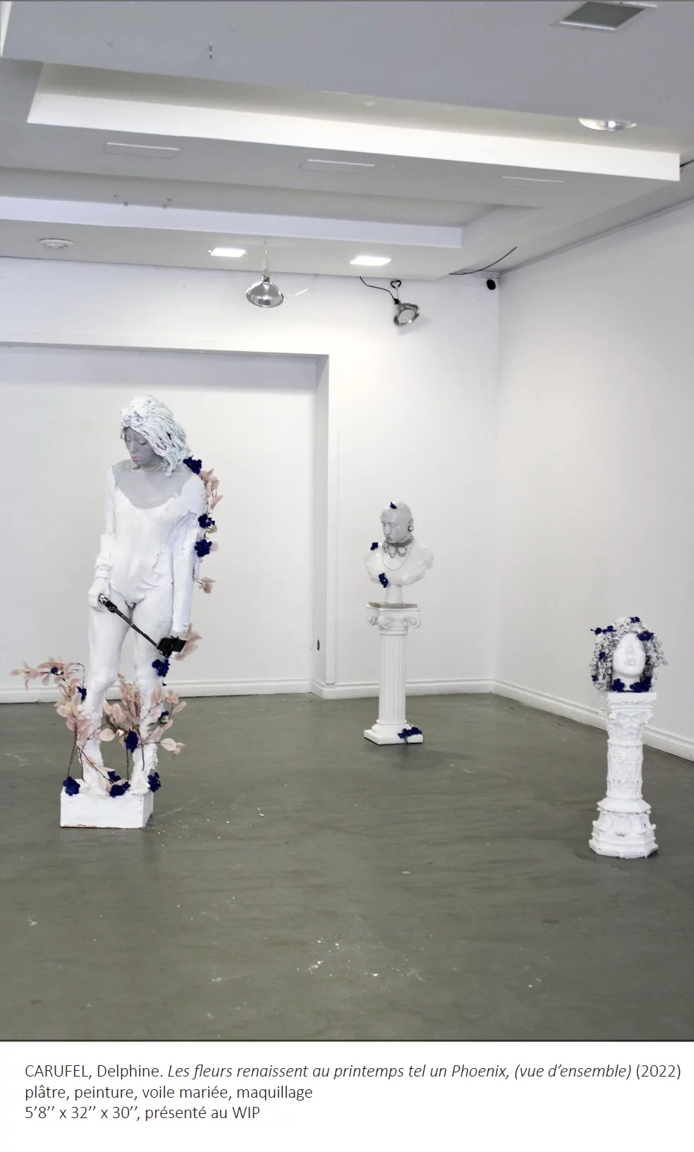 Salle d'exposition avec une sculpture d'un corps humain grandeur nature en plâtre blanc et deux socles blancs avec bustes. Quelques fleurs ornent les trois sculptures.