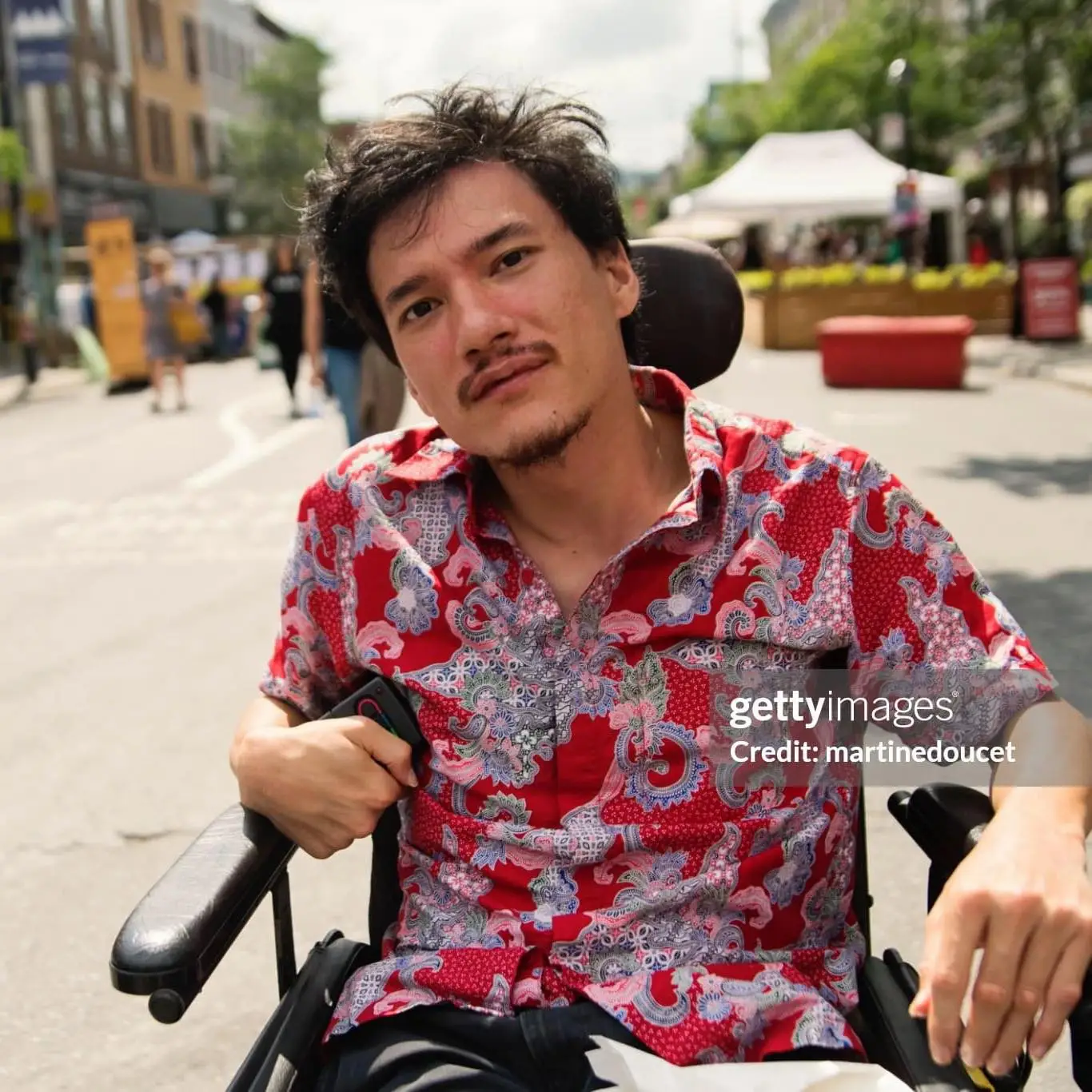 Portrait d'Alexandre Vallerand dans la rue le jour, installé dans son fauteuil roulant électrique, cheveux courts brun, moustache et barbichette courtes brunes, peau olive, chemise rose et bleu fleuris. crédit Getty images Martine Doucet.