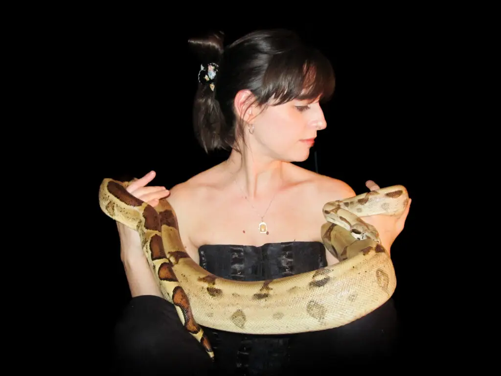 Fond noir Emilie cheveux brun attaché peau pâle épaules nues tient un serpent jaune et noir dans ses mains, elle regarde à sa gauche.