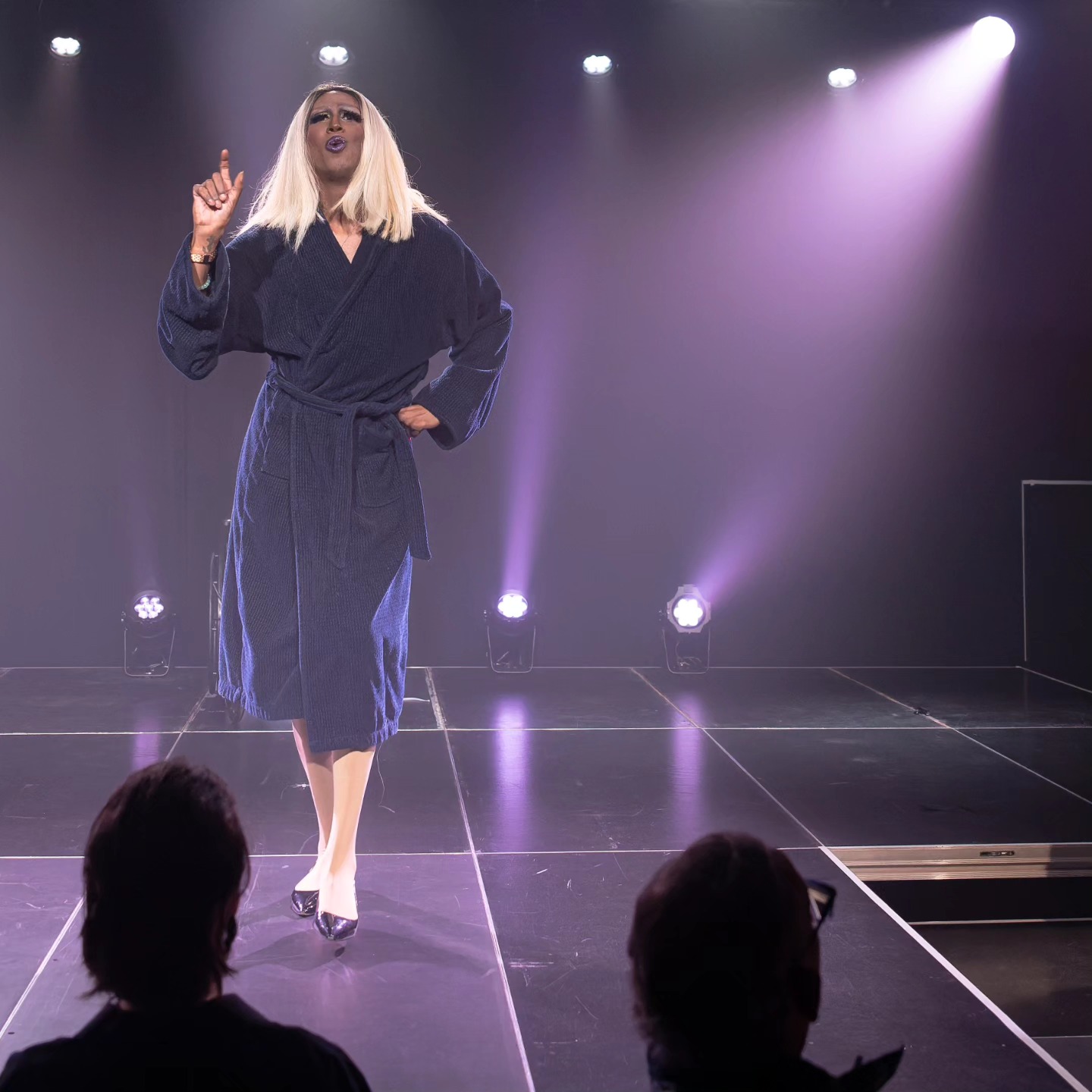 Stella Stone sur scène éclairage mauve, cheveux long blond, peau brune, robe de chambre bleue, souliers noirs, Stella pointe le ciel en marchant vers le public