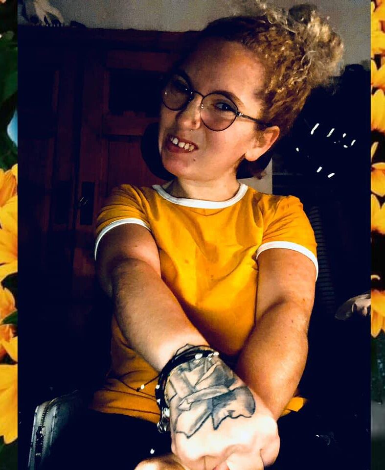 Portrait de l'artiste Joanie-Marika Chouinard deux bande de chaque côté en fleur de tournesol, Joanie t-shirt jaune, lunette de vue regarde l'objectif, les bras en avant croisé laisse voir ses tatoo.