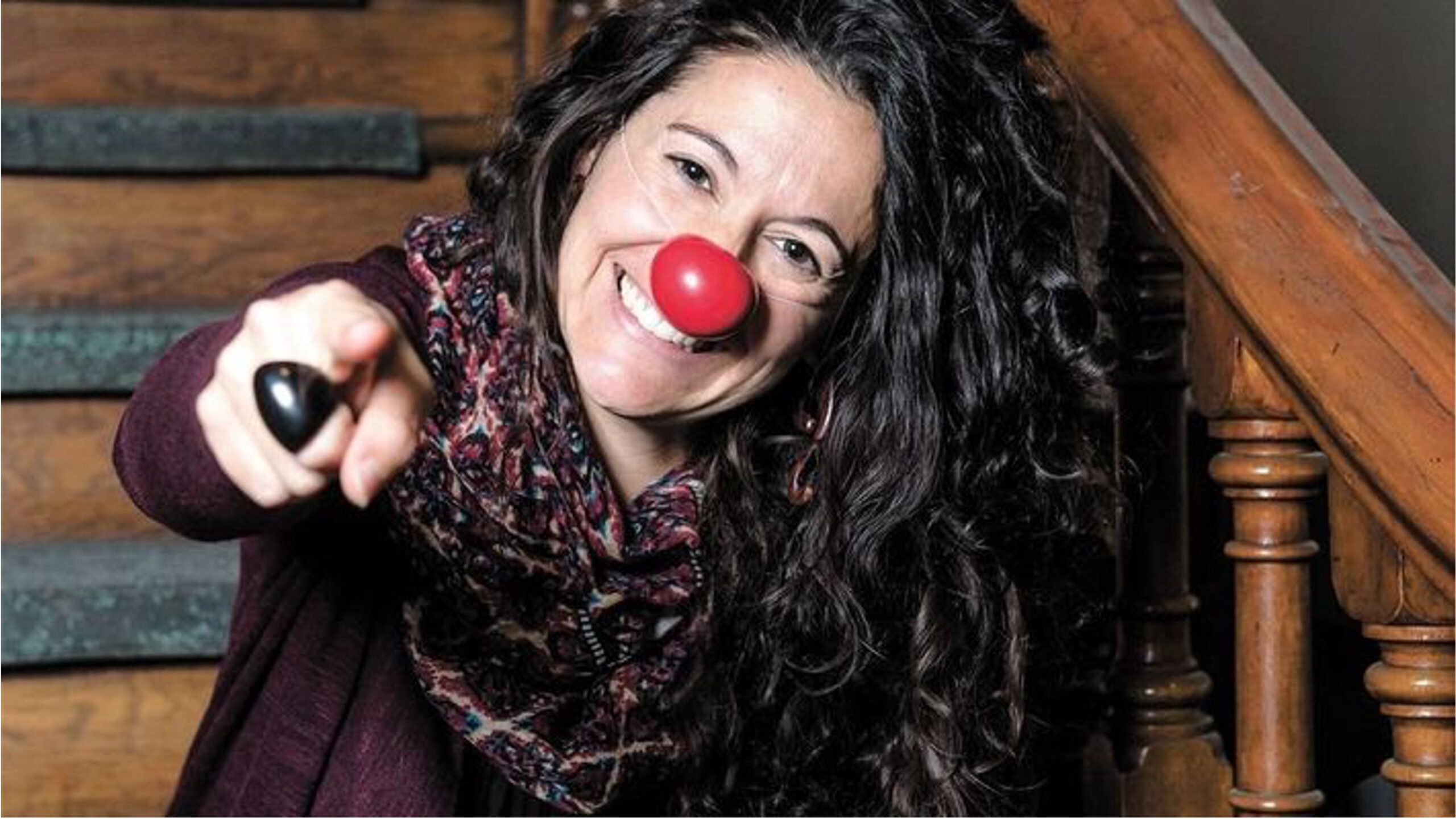 Portrait de Marie-Pierre Petit souriante regard caméra nez rouge elle pointe l'objectif du doigt. Peau blanche longs cheveux brun bouclés.